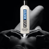 A bottle of "Fuck Water Original Water & Silicone-Based Lubricant" by the brand Fuck Water is showcased against a dark, blurred background with an abstract human figure. The white bottle features eye-catching black and blue text that makes it pop.